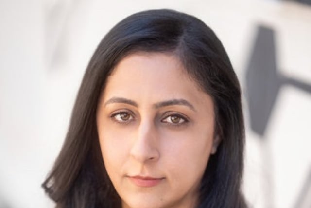 Author Patmeena Sabit gazes at the camera with a serious expression. She has long dark hair and wears a black top.