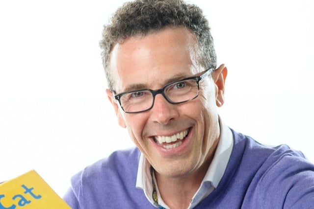 Eric Litwin. A white man with glasses and short dark hair. He is looking into the camera and wearing a blue-purple sweater over a blue collared shirt and holding open a copy of Pete the Cat: I Love My White Shoes.