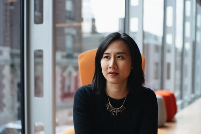 Celeste Ng sitting in an orange chair. Her hands are clasped in her lap and she is wearing a black shirt and black pants with a gold necklace.