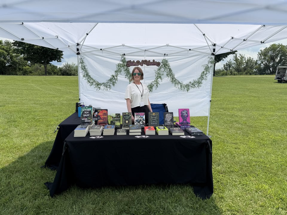 Outdoor white canopy with a garland backdrop and wooden 'Stories of Wonder' sign. There is a table full of colorful books in the foreground.