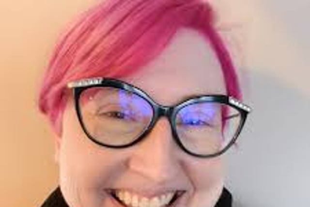 a headshot of robyn bachar, a pink haired white woman with oversized cat eye glasses. she is smiling into the camera and wearing a black shirt.