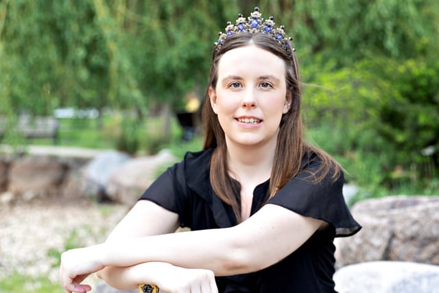 a photo of the author kris julian, a woman with a tiara on her head, leaning on a sword, with a neutral facial expression.